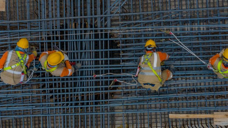 Four construction workers wearing safety gear are working on a large grid of rebar at a construction site